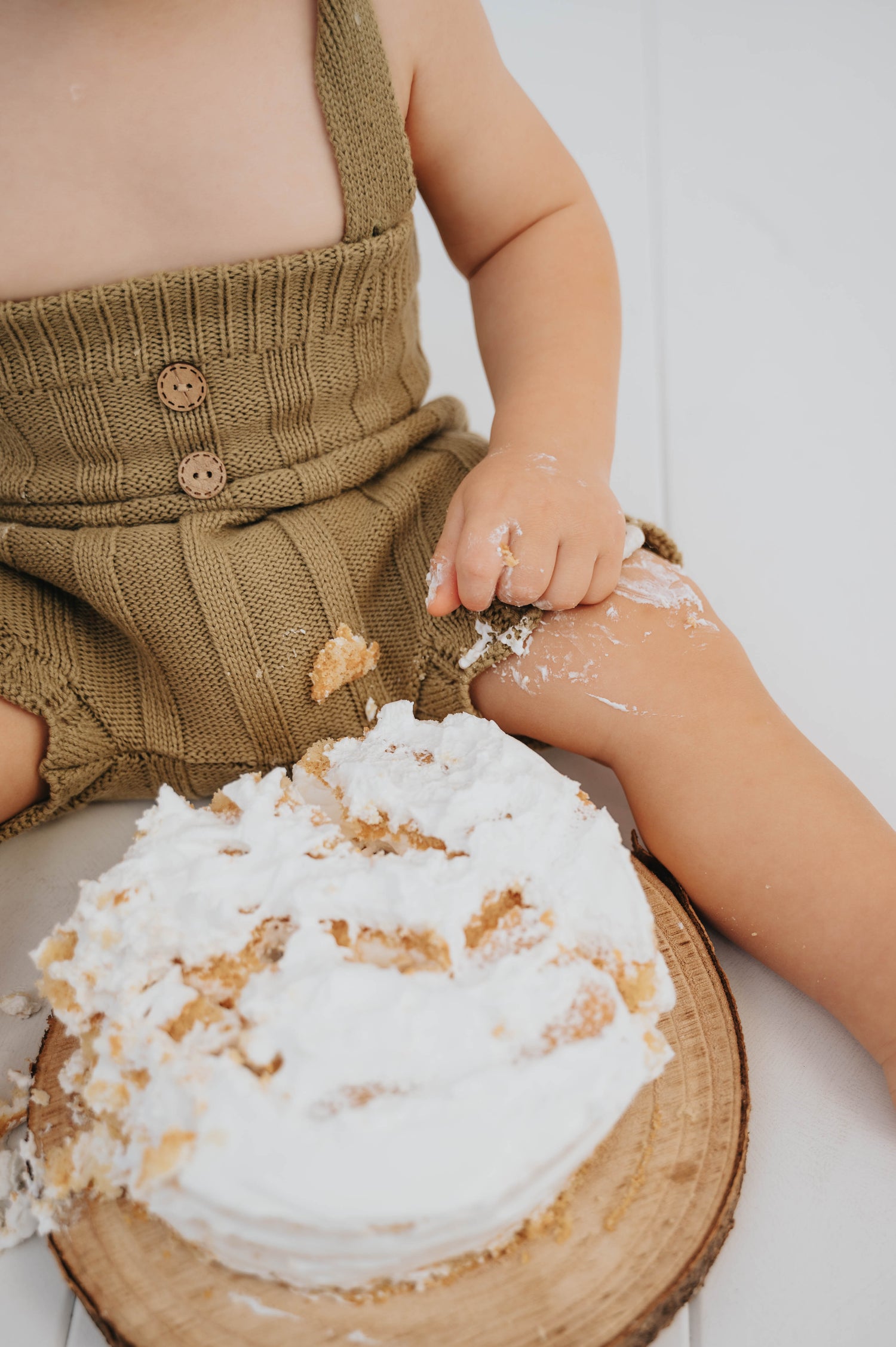Baby wearing knitted brown hlače za otroke sitting next to a messy cake on wooden board