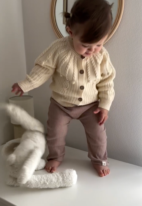 Toddler wearing cozy beige sweater and brown otroške pajkice standing on white surface playing with plush toy