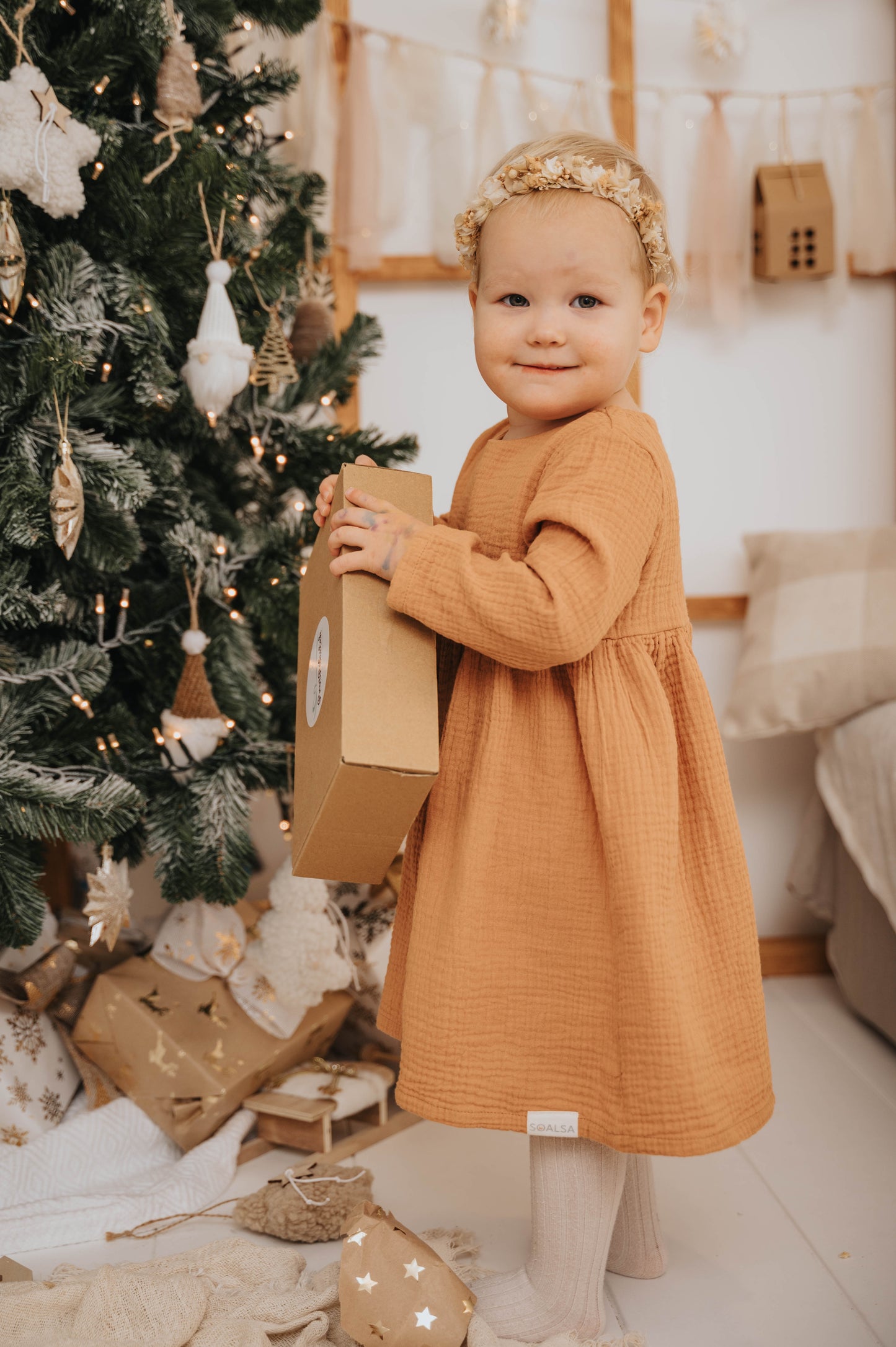 Toddler girl in a brown dress holding a gift near decorated Christmas tree Božična obleka za deklice