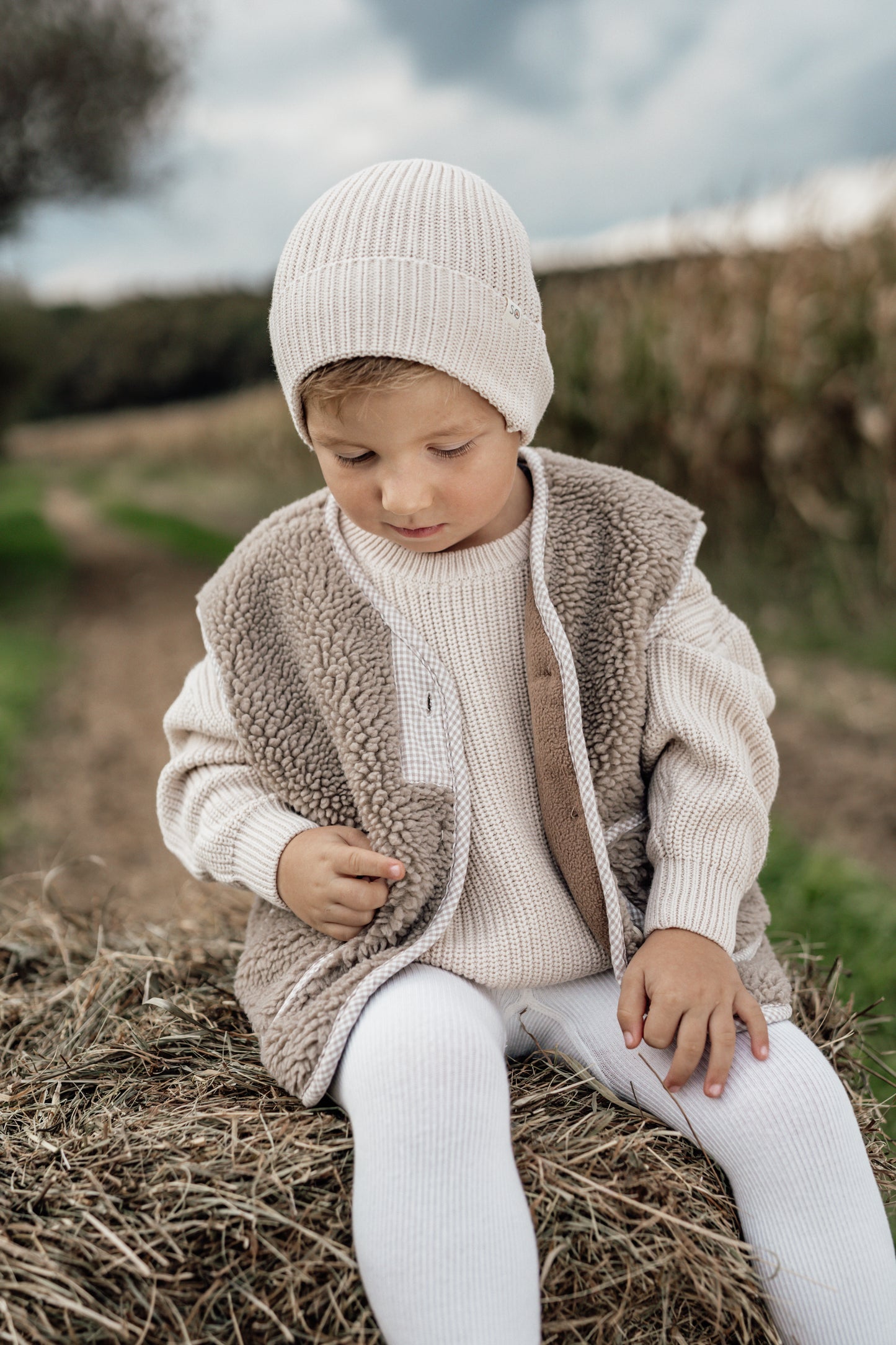 Child wearing warm beige božični puloverji za otroke and matching knit hat sitting on hay