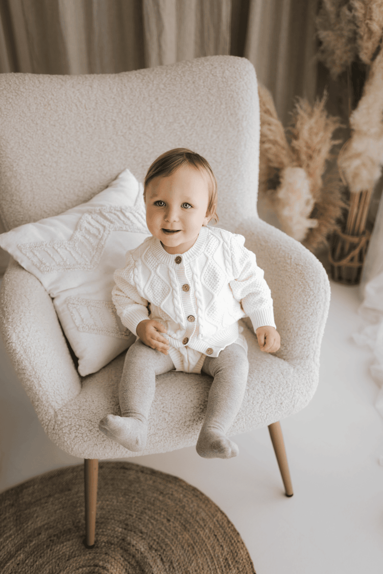 Smiling baby sitting in a cozy chair wearing a white knitted outfit Zimski komplet za novorojenčka for winter