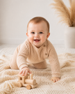 Baby in a beige knitted sweater playing with a wooden toy on a soft surface.