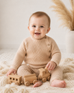 Baby in a beige knit outfit playing with a wooden toy train on a soft surface.