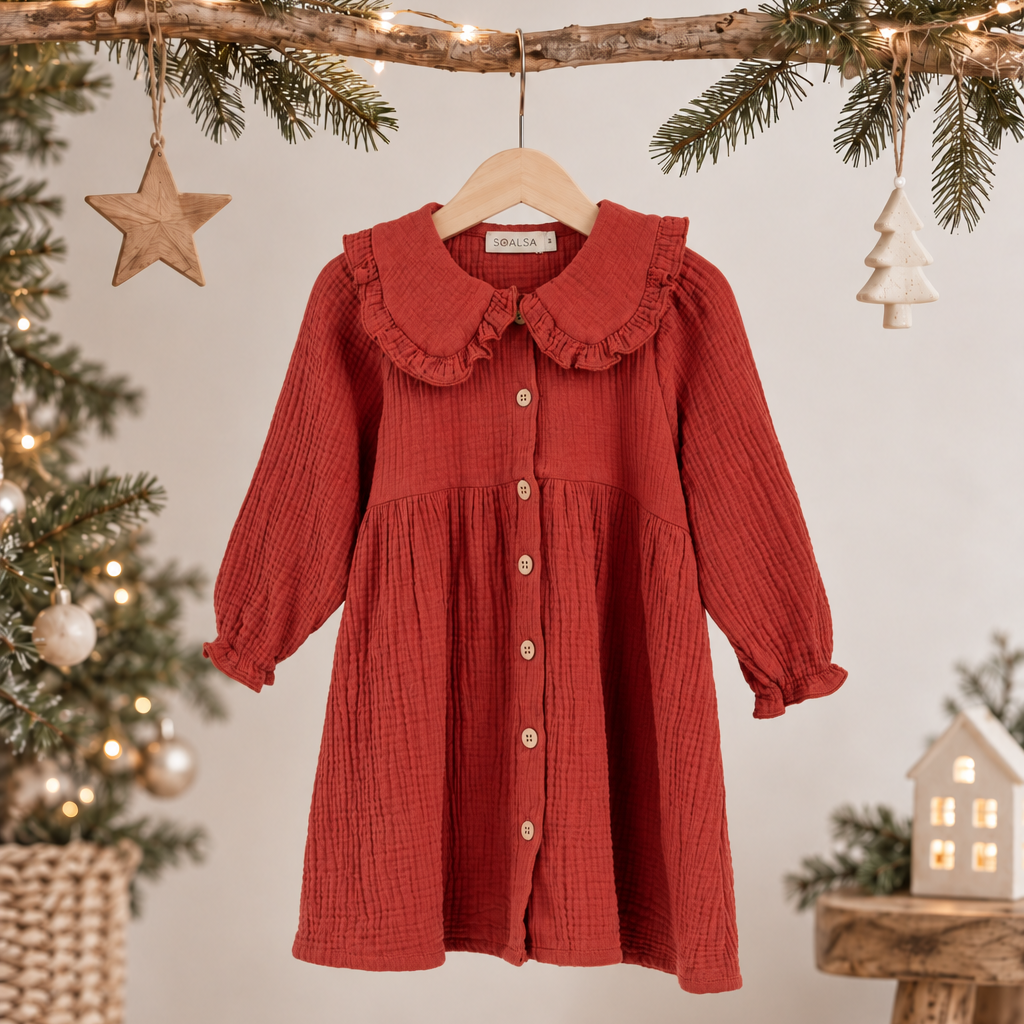 A rust red crinkle-textured cotton dress for toddlers, featuring a large ruffled Peter Pan collar and button-front closure, hanging on a wooden hanger from a decorated Christmas tree branch.