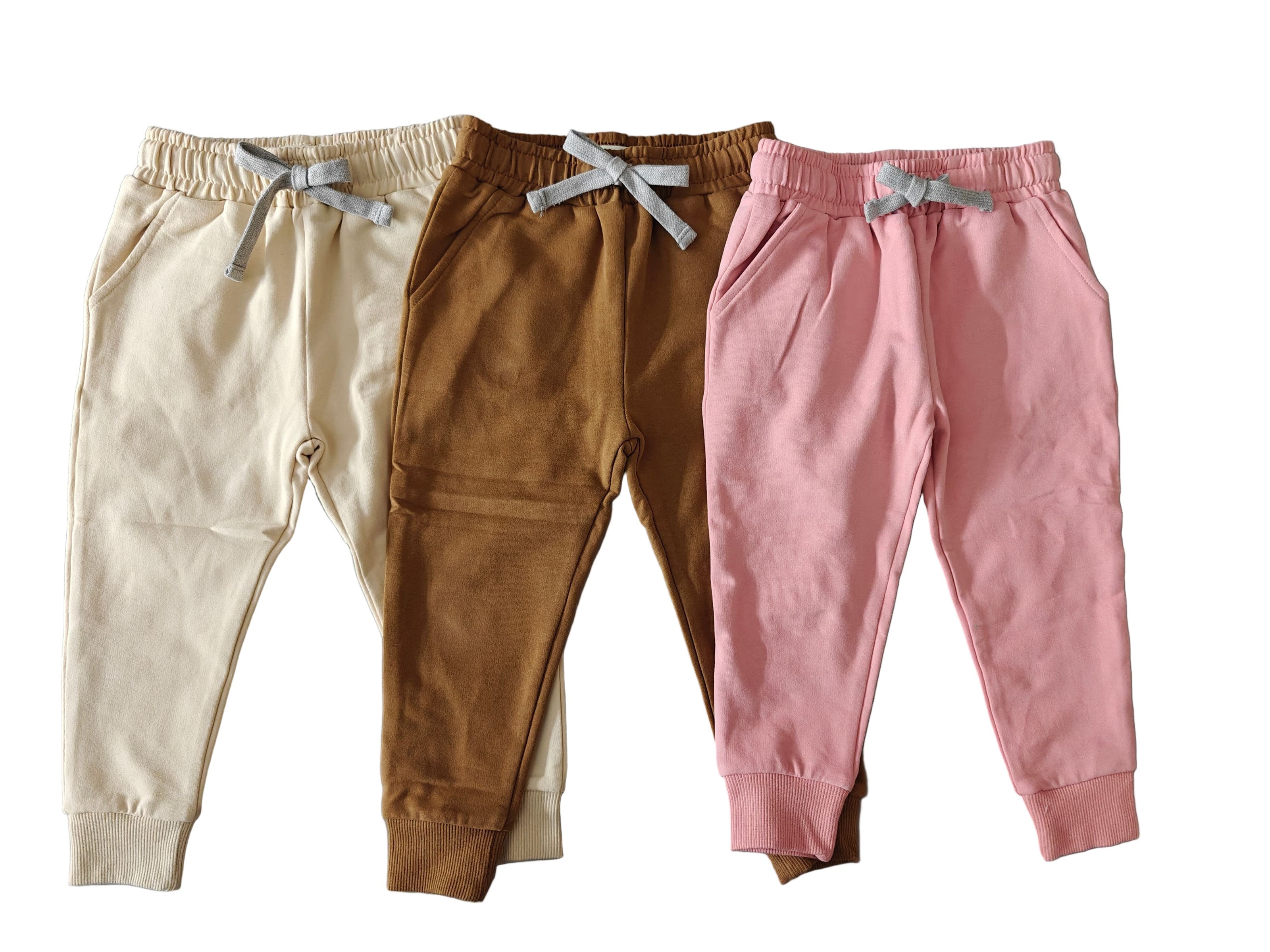 Three pairs of children's jogger pants in beige, brown, and pink on a gray surface.