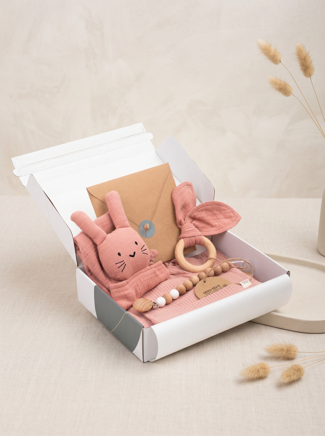 Pink plush bunny toy with a teething ring in an open gift box on a light beige background.