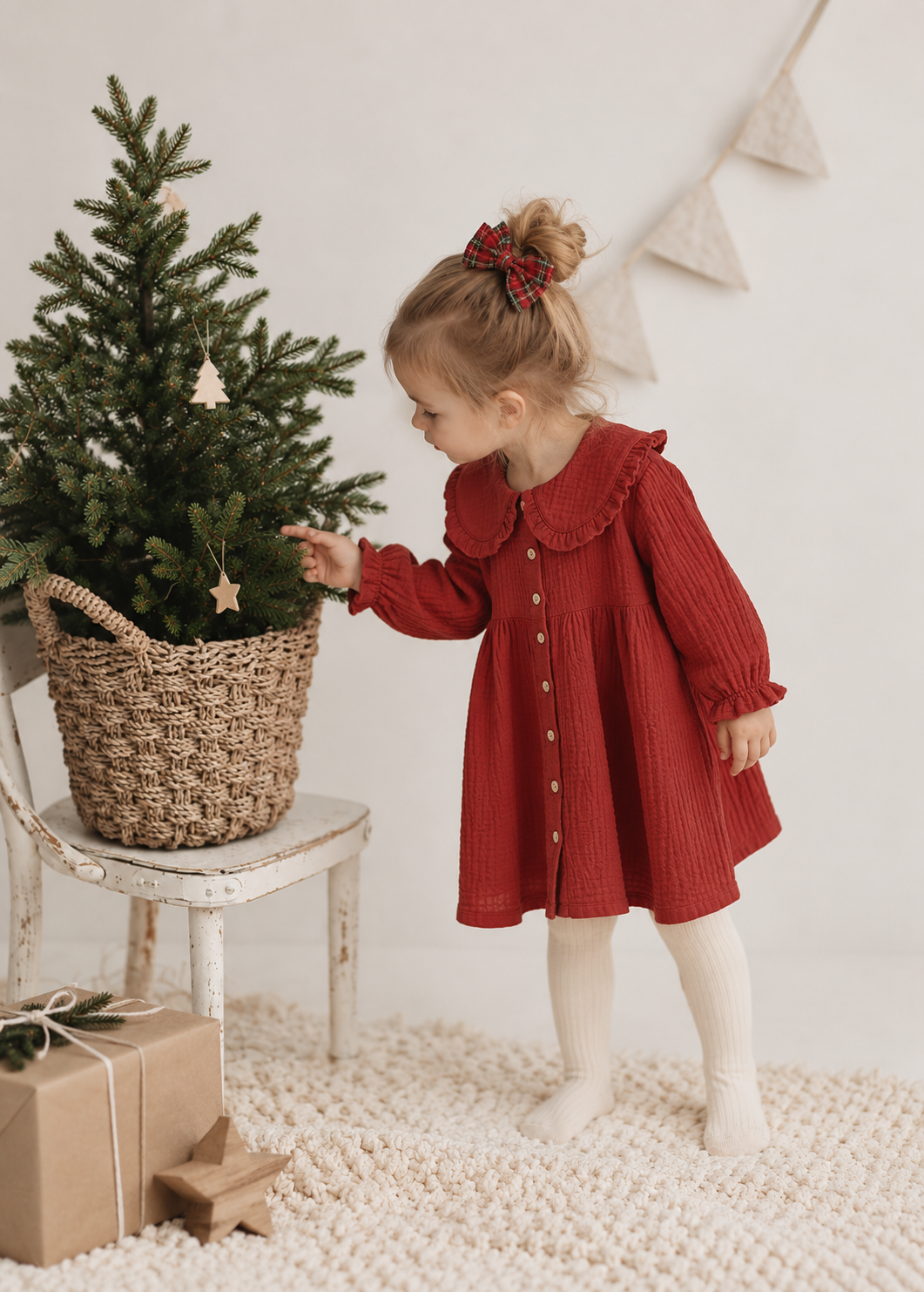 A young girl wearing a red ribbed long-sleeve dress with a Peter Pan collar and white tights stands next to a potted Christmas tree in a styled indoor setting.