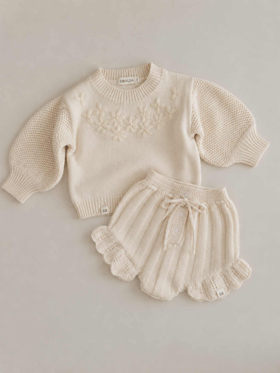 Cream-colored knitted baby outfit with a sweater and shorts on a light gray background