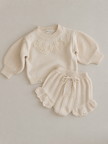 Cream-colored knitted baby outfit with a sweater and shorts on a light gray background