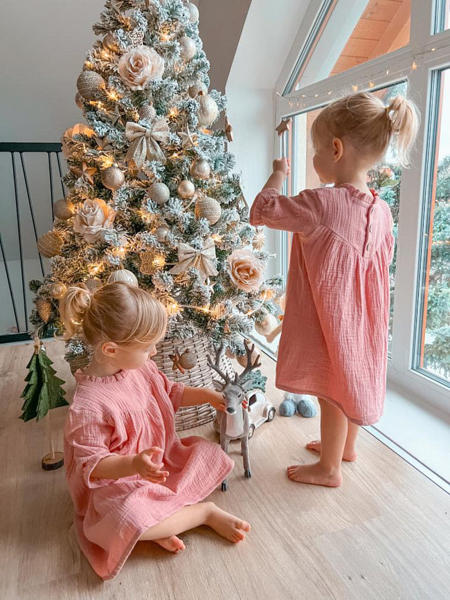 Two toddlers wearing pink Božična obleka za dojenčke dresses near a decorated Christmas tree indoors