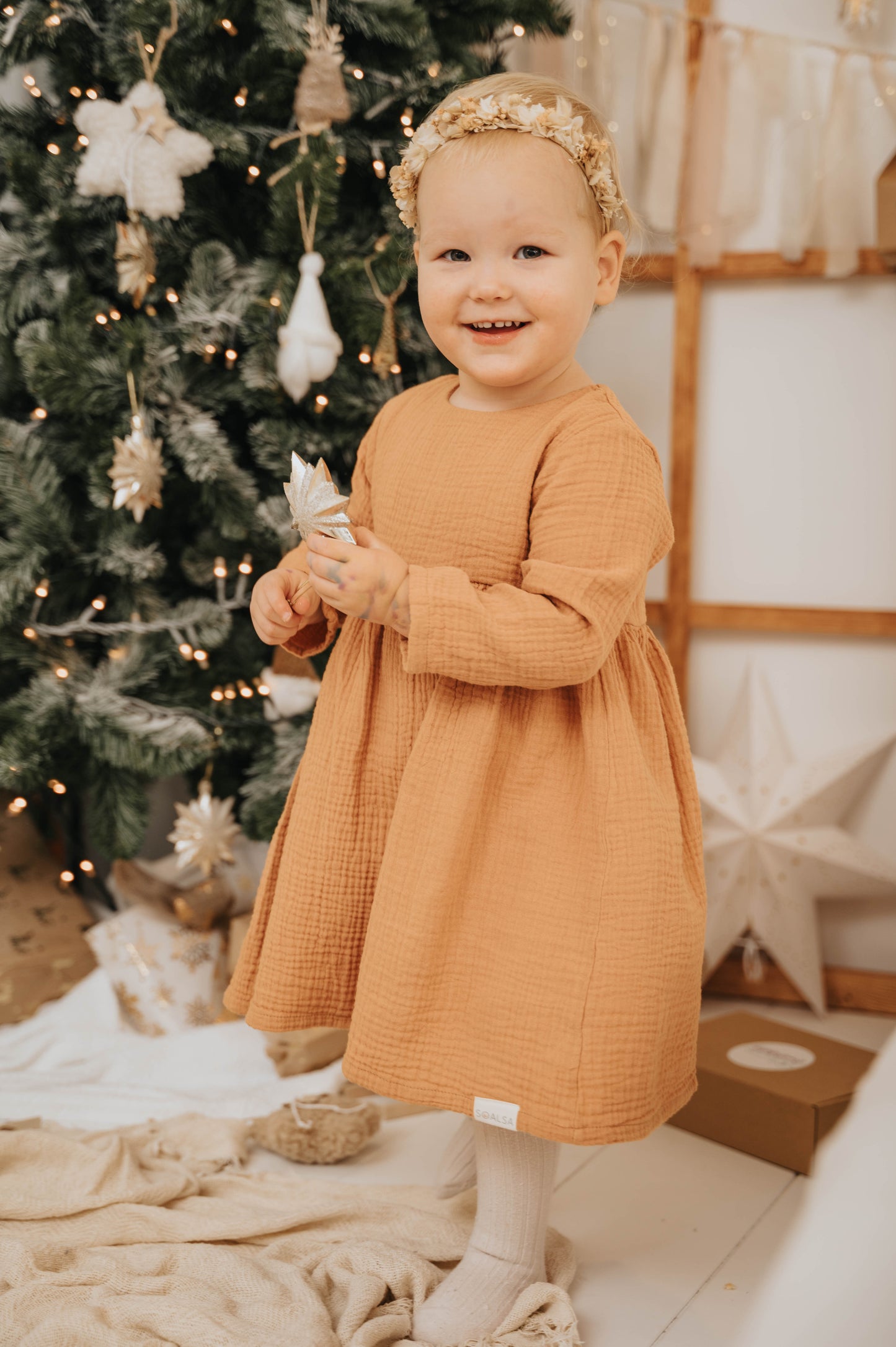 Toddler girl wearing a mustard dress and floral headband standing by Christmas tree Božična obleka za deklice