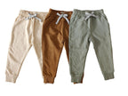 Three pairs of children's jogger pants in beige, brown, and green on a gray background.