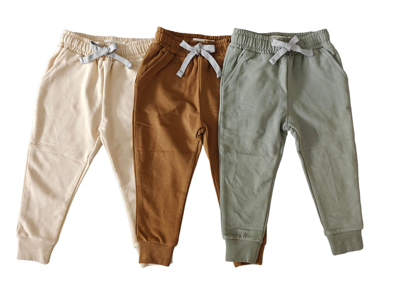 Three pairs of children's jogger pants in beige, brown, and green on a gray background.