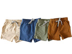 Four pairs of children's shorts in beige, blue, green, and brown on a gray floor.
