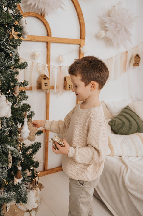 Young boy decorating Christmas tree wearing cozy beige sweater for božični puloverji za otroke