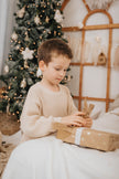 Young boy in cream sweater opening gift near decorated Christmas tree božični puloverji za otroke