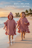Two children wearing pink hooded towels on a beach with text about organic cotton and fast drying.