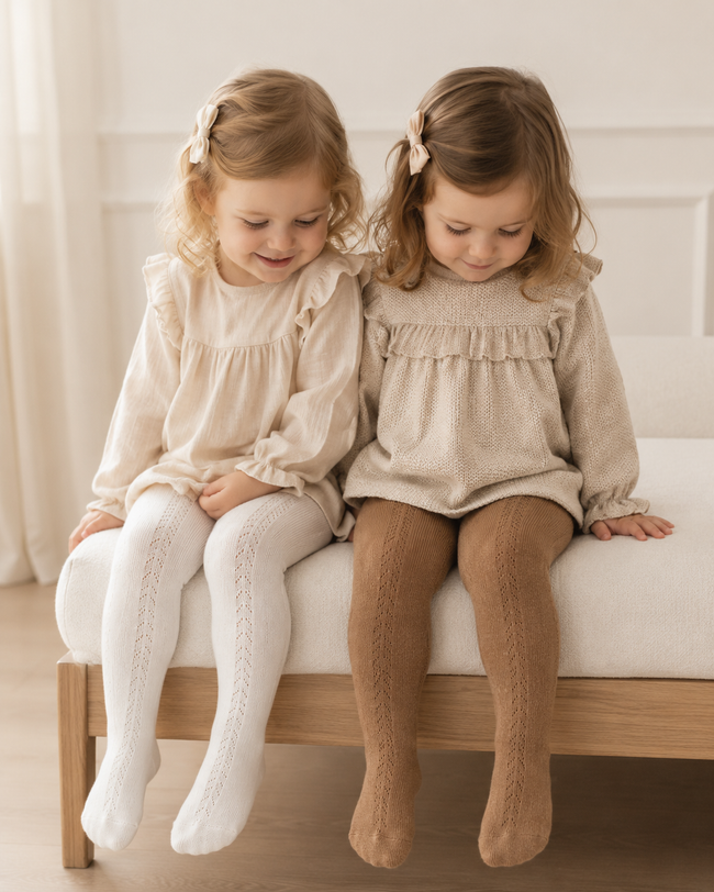 Two young girls sitting side by side on a white cushioned bench, wearing matching long-sleeve ruffle-trimmed tops in cream and beige, paired with white and tan knit tights.